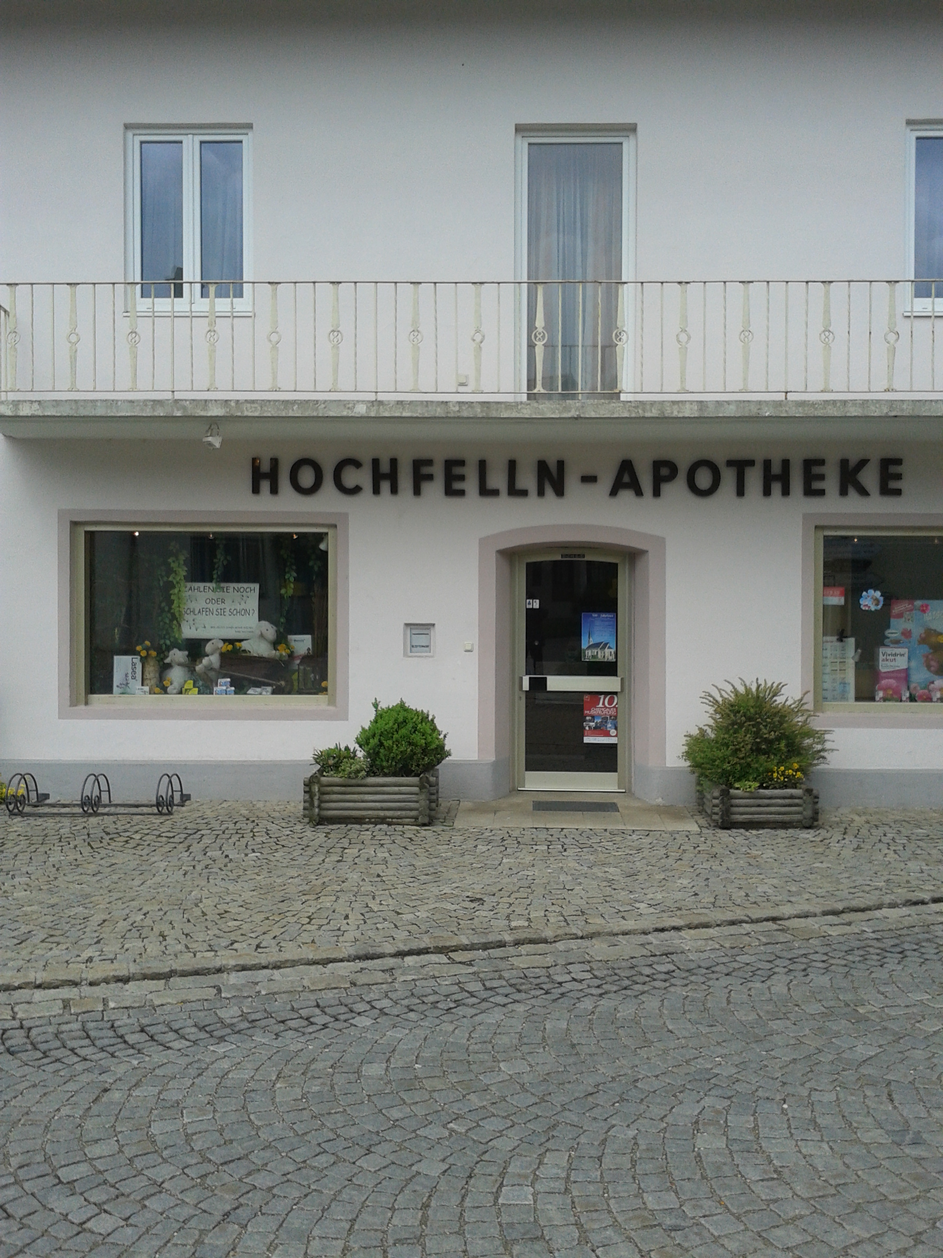 Frontalansicht der Apotheke mit Briefkasten. An der Tür weitere Reklameposter,außerdem ist der Balkon des Hauses zu sehen.